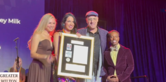 Group of four people on stage posing with a framed award plaque, smiling for the camera; purple backdrop and sign reading 'Greater Wilton Manors' visible.