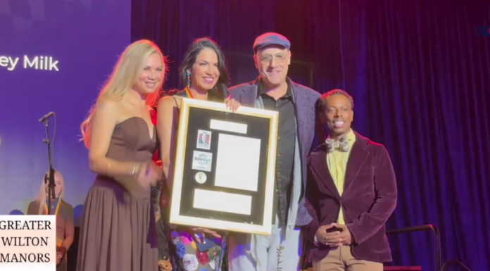 Group of four people on stage posing with a framed award plaque, smiling for the camera; purple backdrop and sign reading 'Greater Wilton Manors' visible.