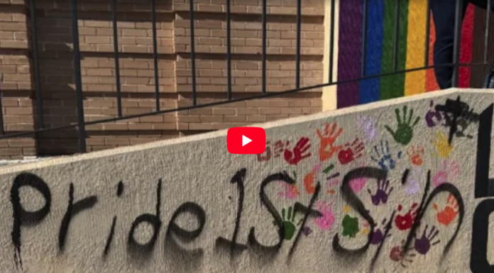 Video thumbnail showing graffiti on a concrete wall: the text 'pride is' with multicolored handprints around it and a red play button overlay in the center.
