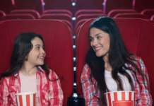 Two smiling girls in red plaid shirts sitting in a movie theater with popcorn tubs, facing each other and talking.