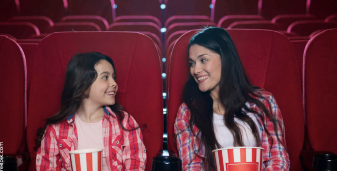 Two smiling girls in red plaid shirts sitting in a movie theater with popcorn tubs, facing each other and talking.