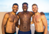 Three shirtless men smiling with arms around each other on a sunny beach by the sea.