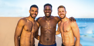 Three shirtless men smiling with arms around each other on a sunny beach by the sea.