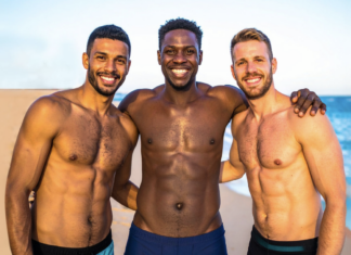 Three shirtless men smiling with arms around each other on a sunny beach by the sea.
