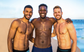 Three shirtless men smiling with arms around each other on a sunny beach by the sea.