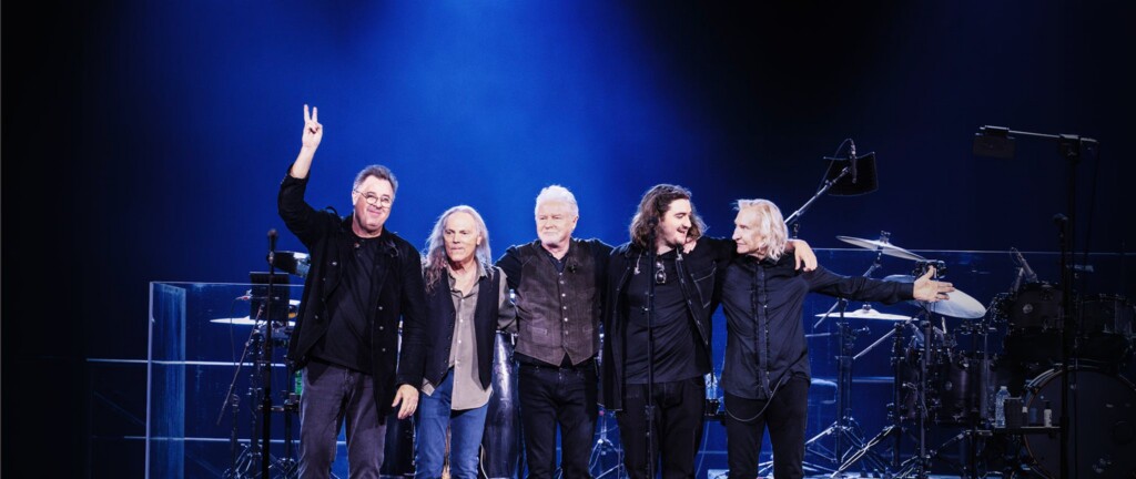 Five male musicians stand together on a stage with arms around each other under blue stage lighting, after a performance.