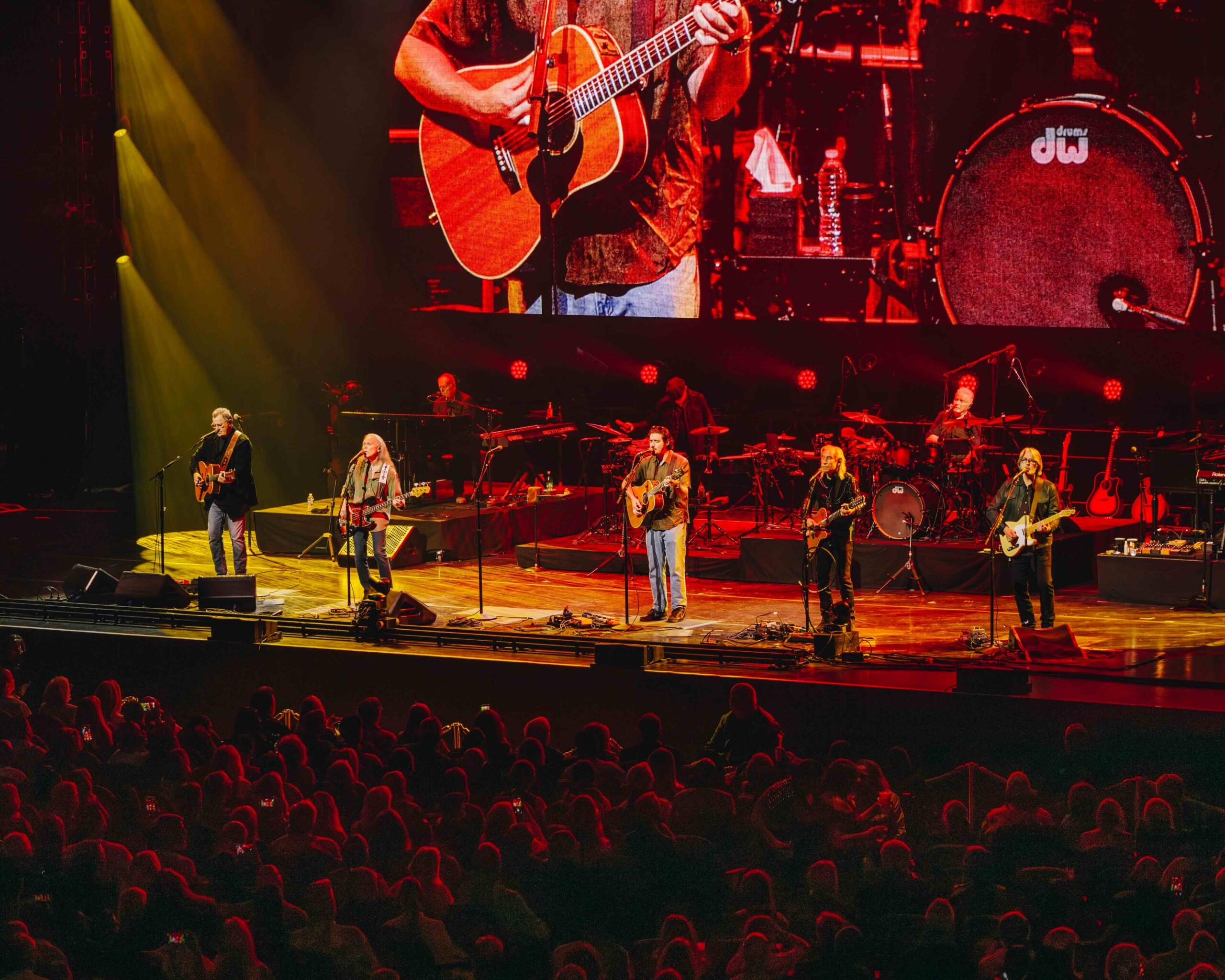 Live concert on a large stage with guitarists and a drummer under red and yellow lighting; audience silhouettes fill the foreground.