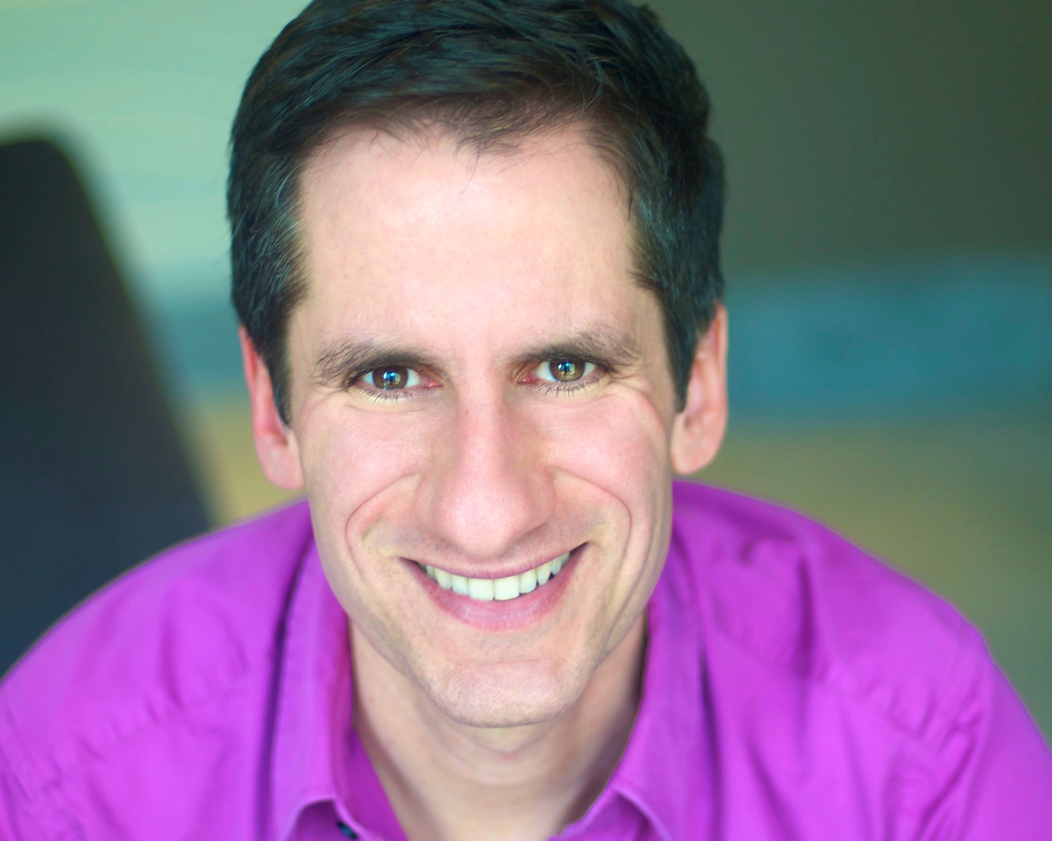 Close-up portrait of a smiling man with short dark hair wearing a magenta shirt, looking at the camera.