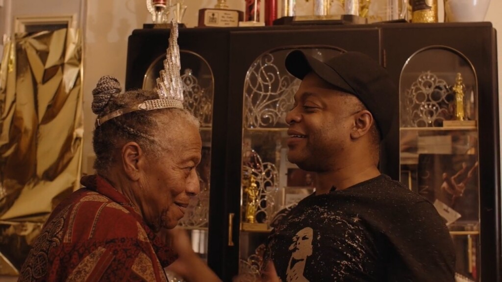 Prime Video Premieres “Pageant Queens: Our Story Behind the Crown” | Hotspots Magazine Hotspots Magazine Older man wearing a silver crown smiles at a younger man in a black cap; trophies on display behind them.
