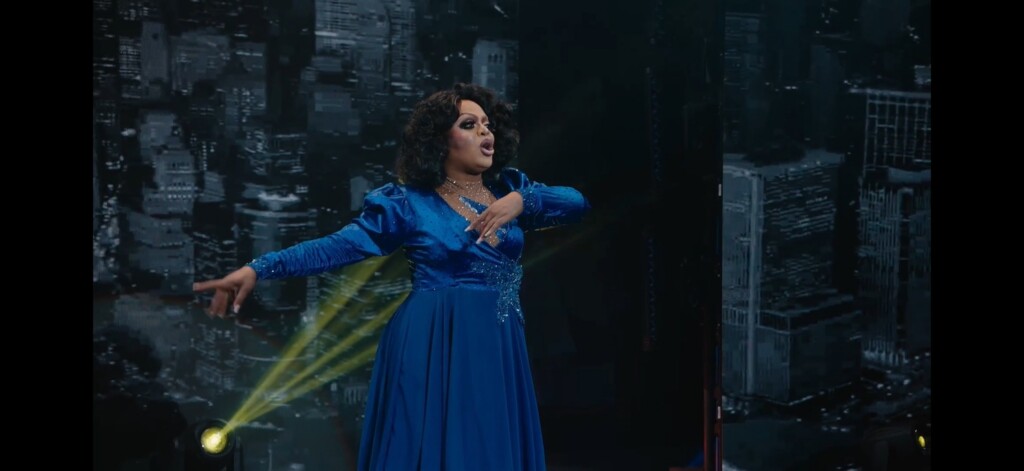 Prime Video Premieres “Pageant Queens: Our Story Behind the Crown” | Hotspots Magazine Hotspots Magazine Drag performer in a sparkling blue gown mid-performance on a stage, with a yellow spotlight and a city skyline backdrop.