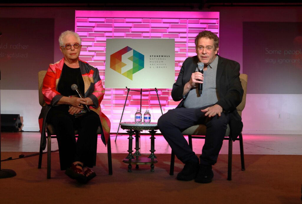 Two panelists sit on stage with microphones, pink backdrop, and a geometric logo sign between them.