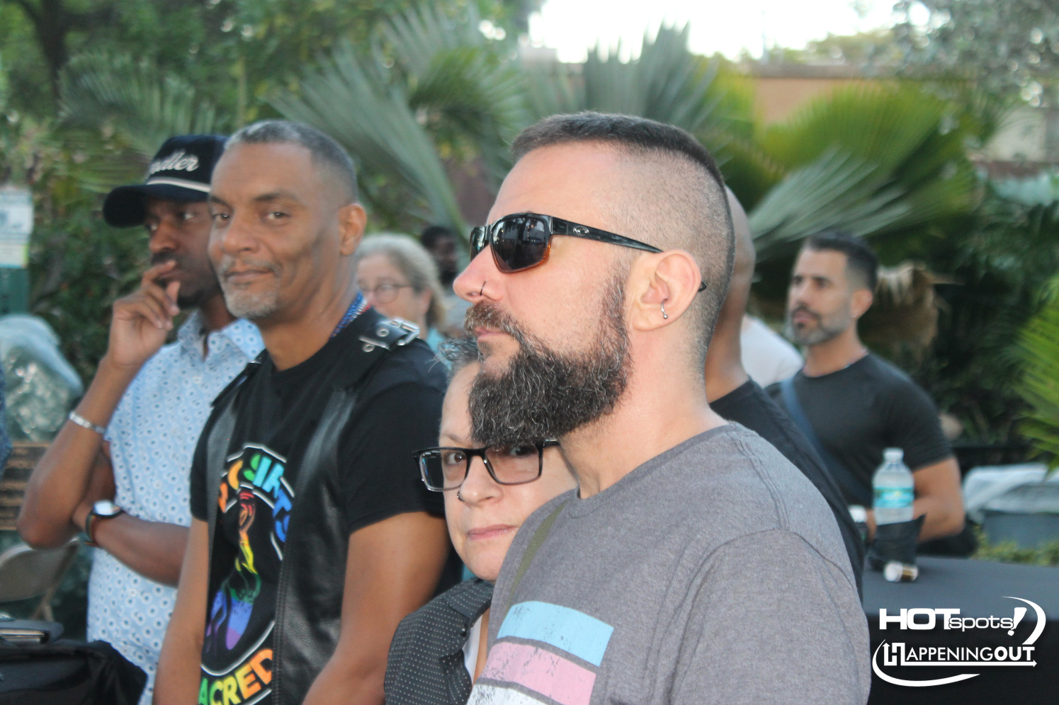 Bearded man with sunglasses and gray t‑shirt in the foreground at an outdoor gathering.