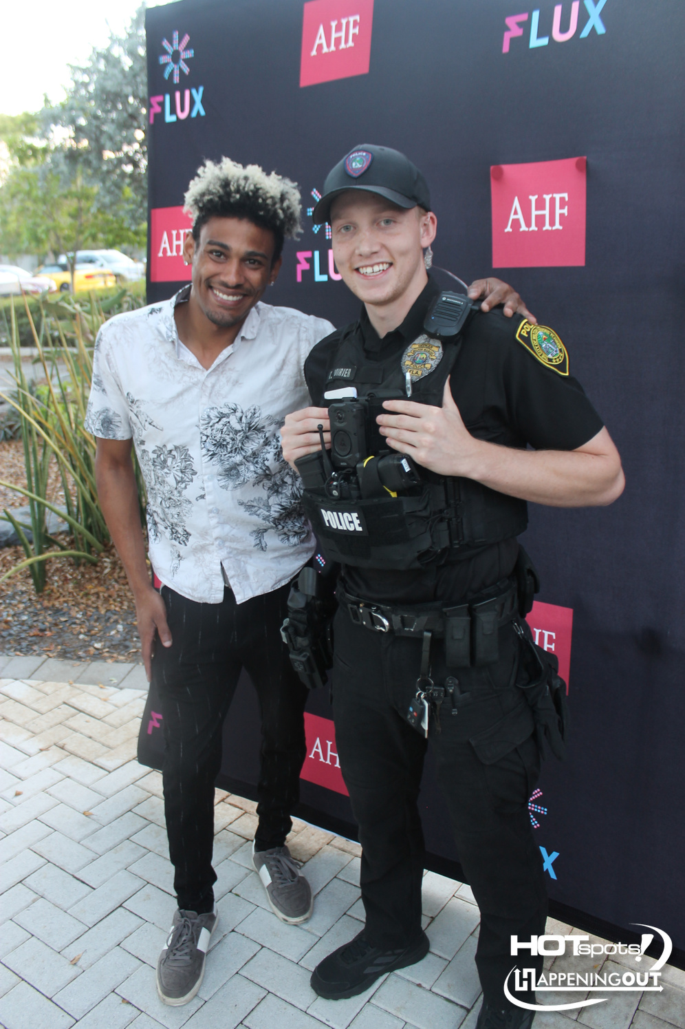 Two men pose for a photo at an event: a smiling civilian in a white floral shirt and a uniformed officer in tactical gear with a camera, standing in front of a branded backdrop.