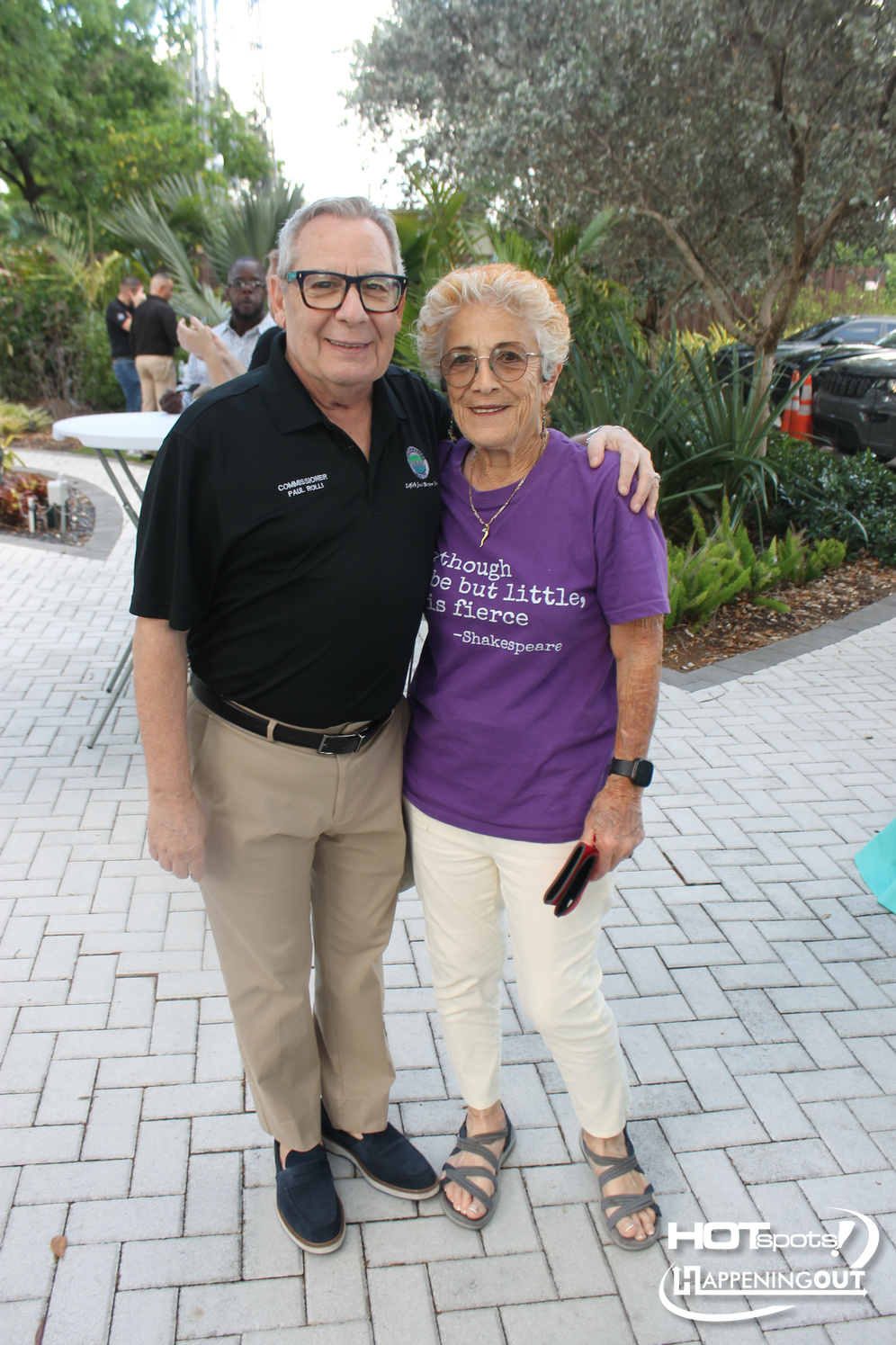 Two smiling older adults pose together outdoors at a garden event; man in a black polo, woman in a purple Shakespeare quote shirt, arms around each other.