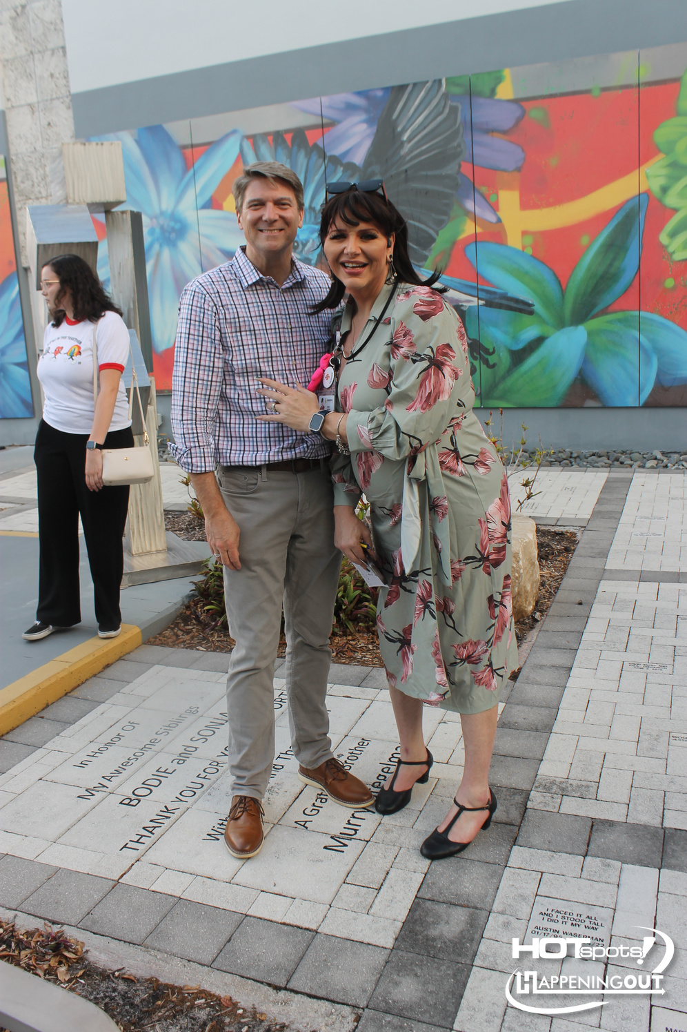 Smiling couple posing for a photo outdoors in front of a bright mural; a third person stands in the background on the left.