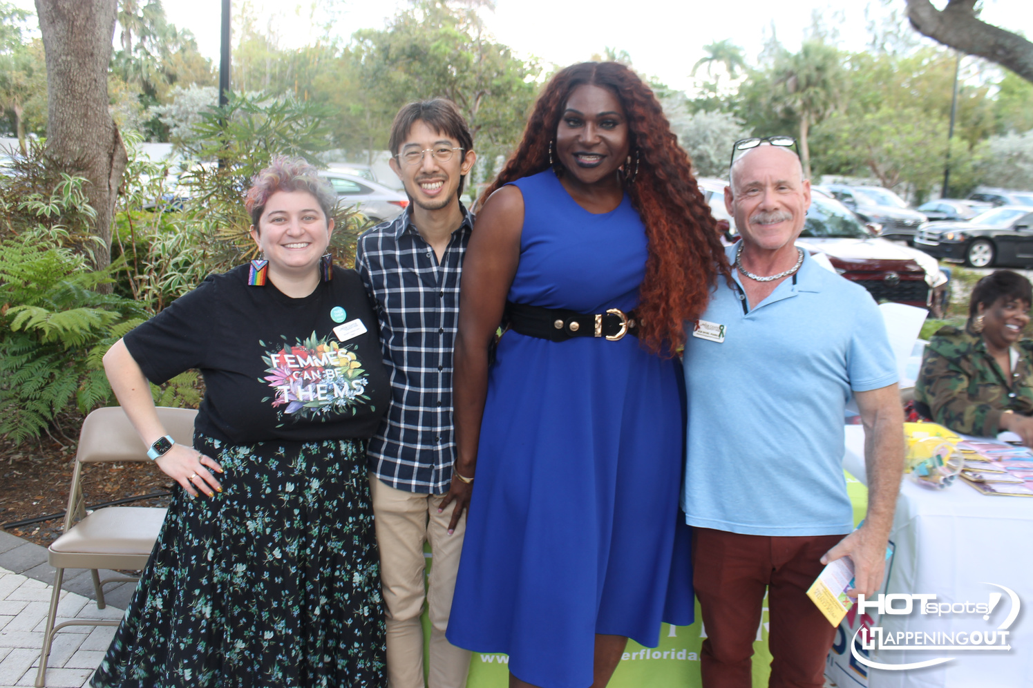 Group portrait of four smiling individuals outdoors at an event, with one person in a blue dress center-right and others in casual attire nearby.