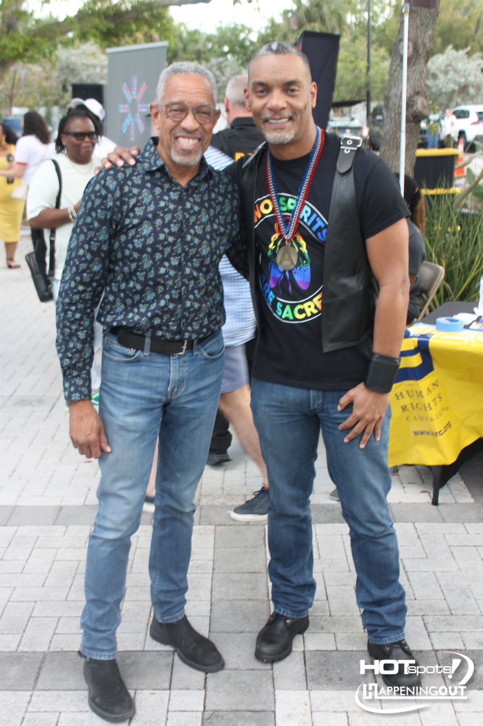 Two men pose together at an outdoor event, smiling for the camera. Left man wears a blue patterned shirt and jeans; right man wears a black T-shirt with a colorful graphic, a leather vest, and beaded necklaces.