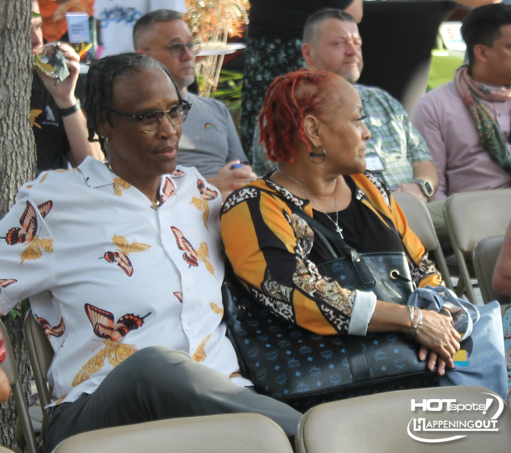 Two adults sit side by side in folding chairs at an outdoor event, a man in a butterfly-patterned shirt and a woman with red hair wearing a black and orange top.