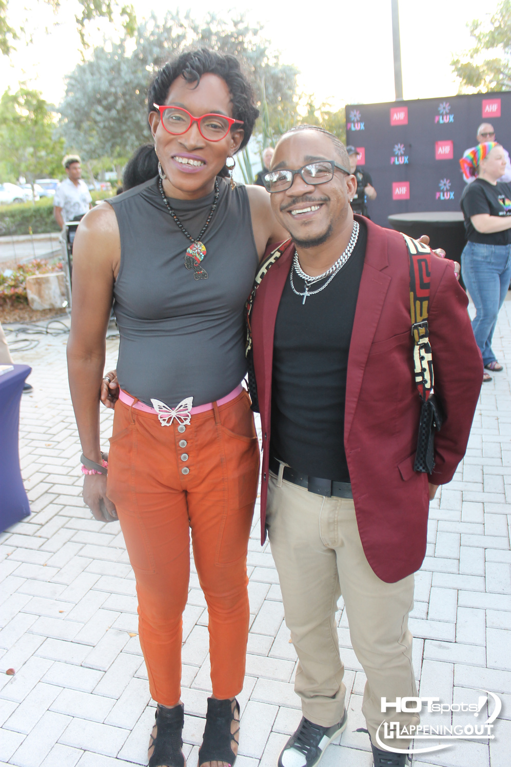 Two stylish friends pose for a photo at an outdoor event, smiling at the camera; the woman wears red glasses, a gray sleeveless top, orange pants with a butterfly belt, and black sandals, while the man wears a maroon blazer, black shirt, beige pants, and multiple necklaces.