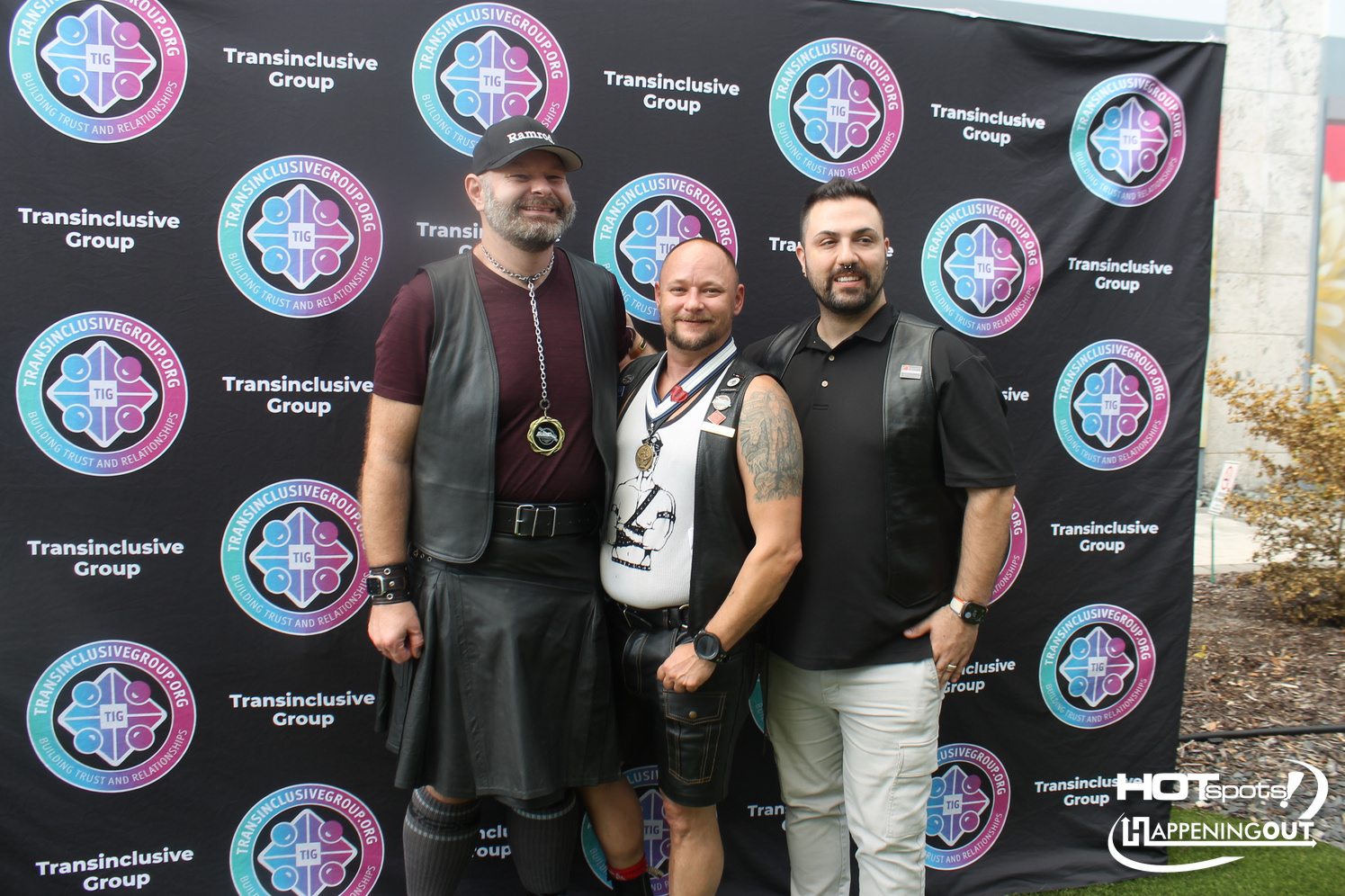 Three men in leather outfits pose together in front of a black step-and-repeat banner featuring the Transinclusive Group logo.