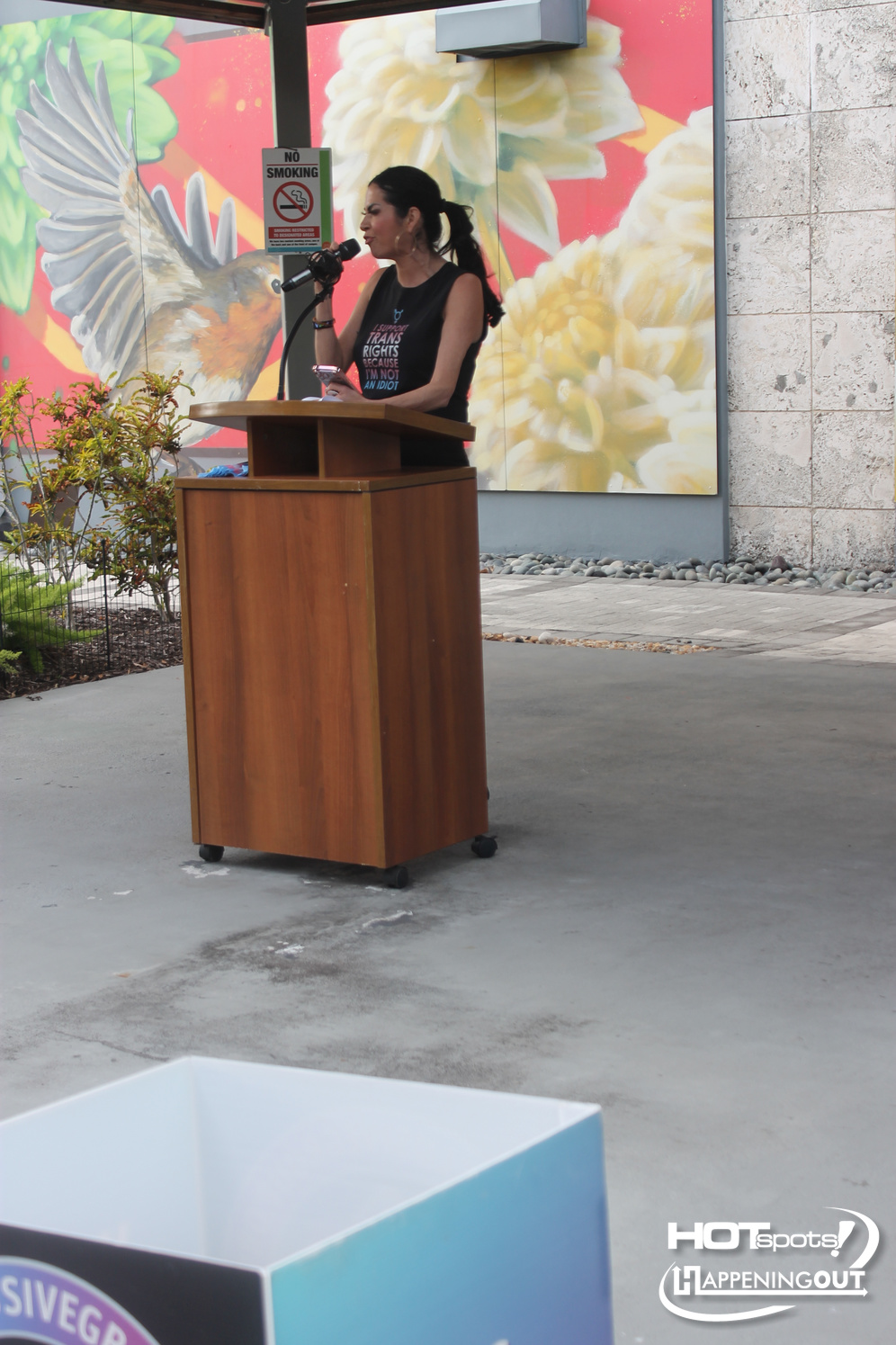 Woman speaks into a microphone at a wooden podium outdoors, with a colorful mural and a No Smoking sign in the background.