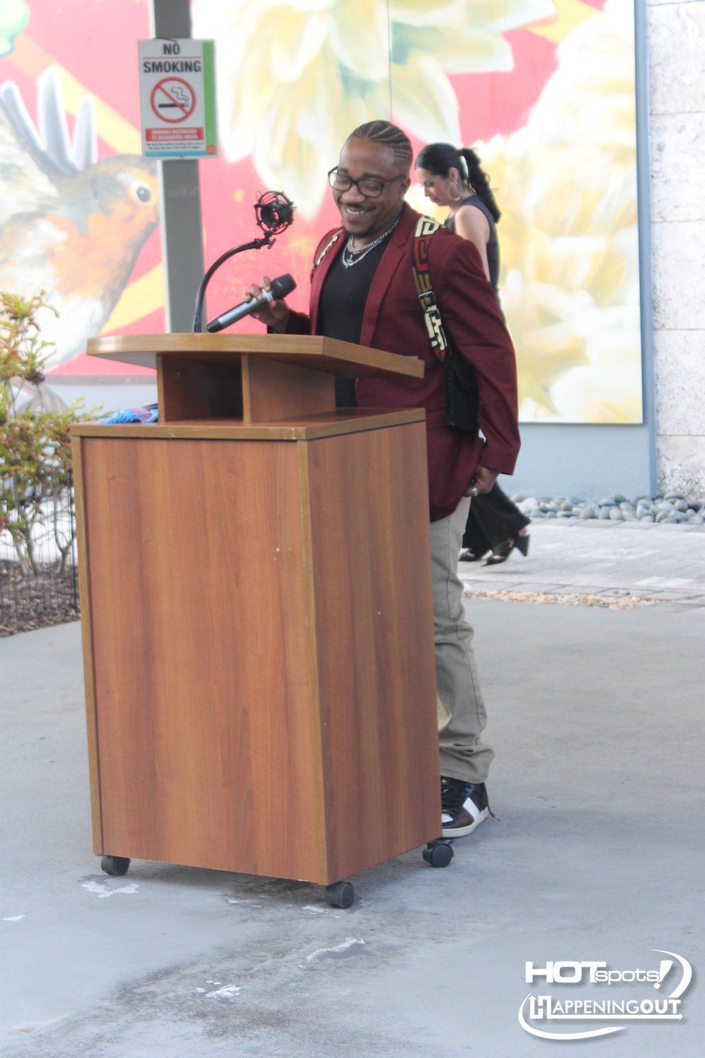 Man in a maroon blazer speaks into a microphone at a wooden podium outdoors, with a colorful mural behind him and a no-smoking sign nearby.