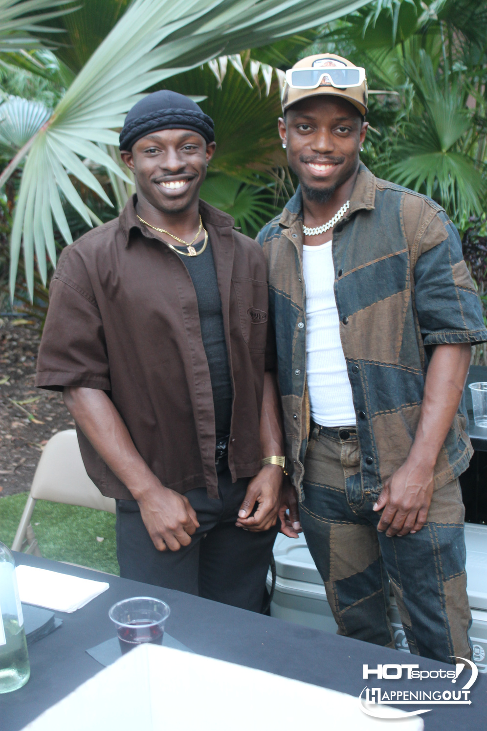 Two men smiling and posing at an outdoor garden party, standing beside a table with drinks, tropical plants in the background.