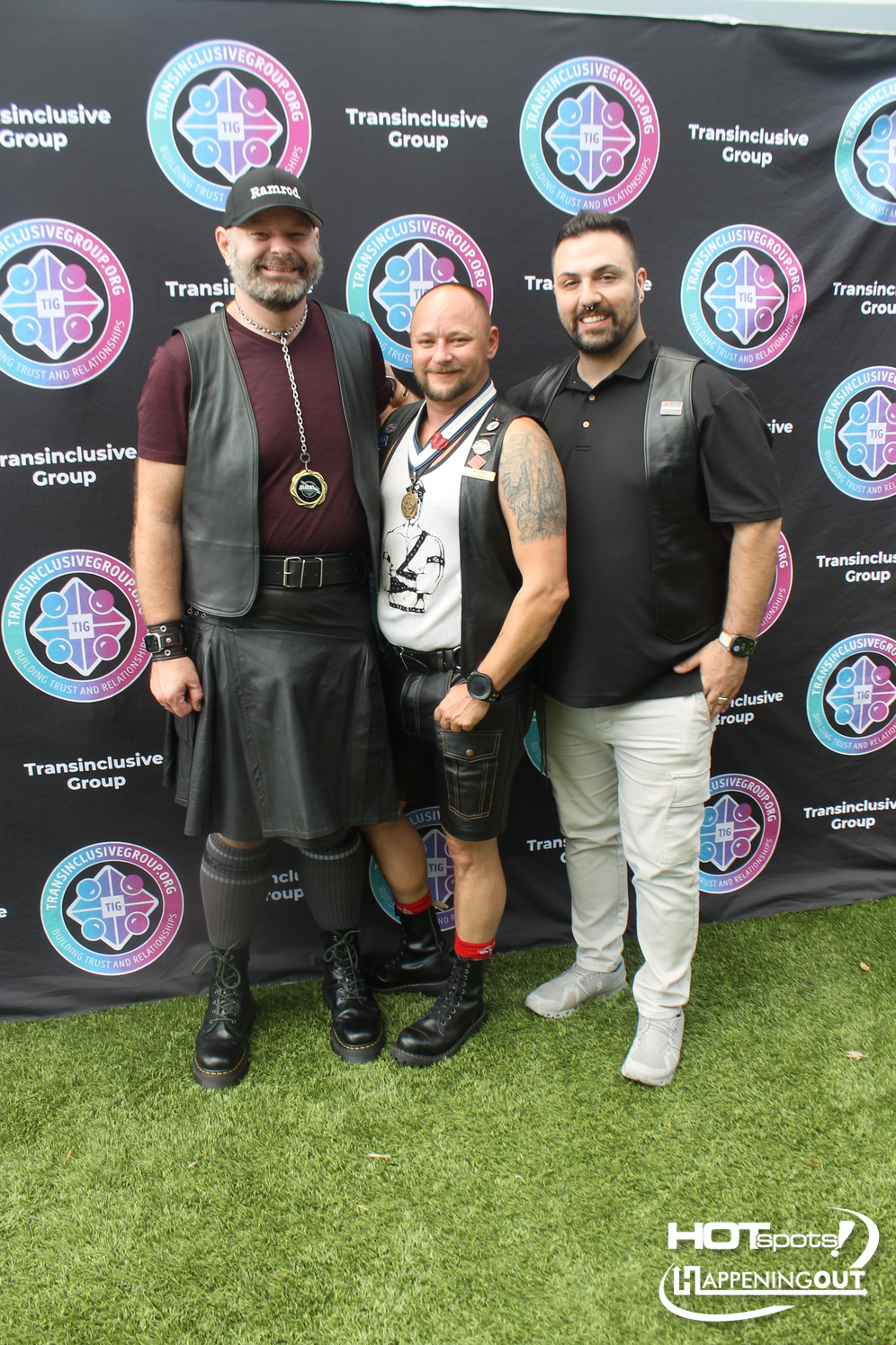Three men in leather outfits pose and smile at an outdoor event, standing in front of a black backdrop covered with Transinclusive Group logos and badges on their chests and necks.