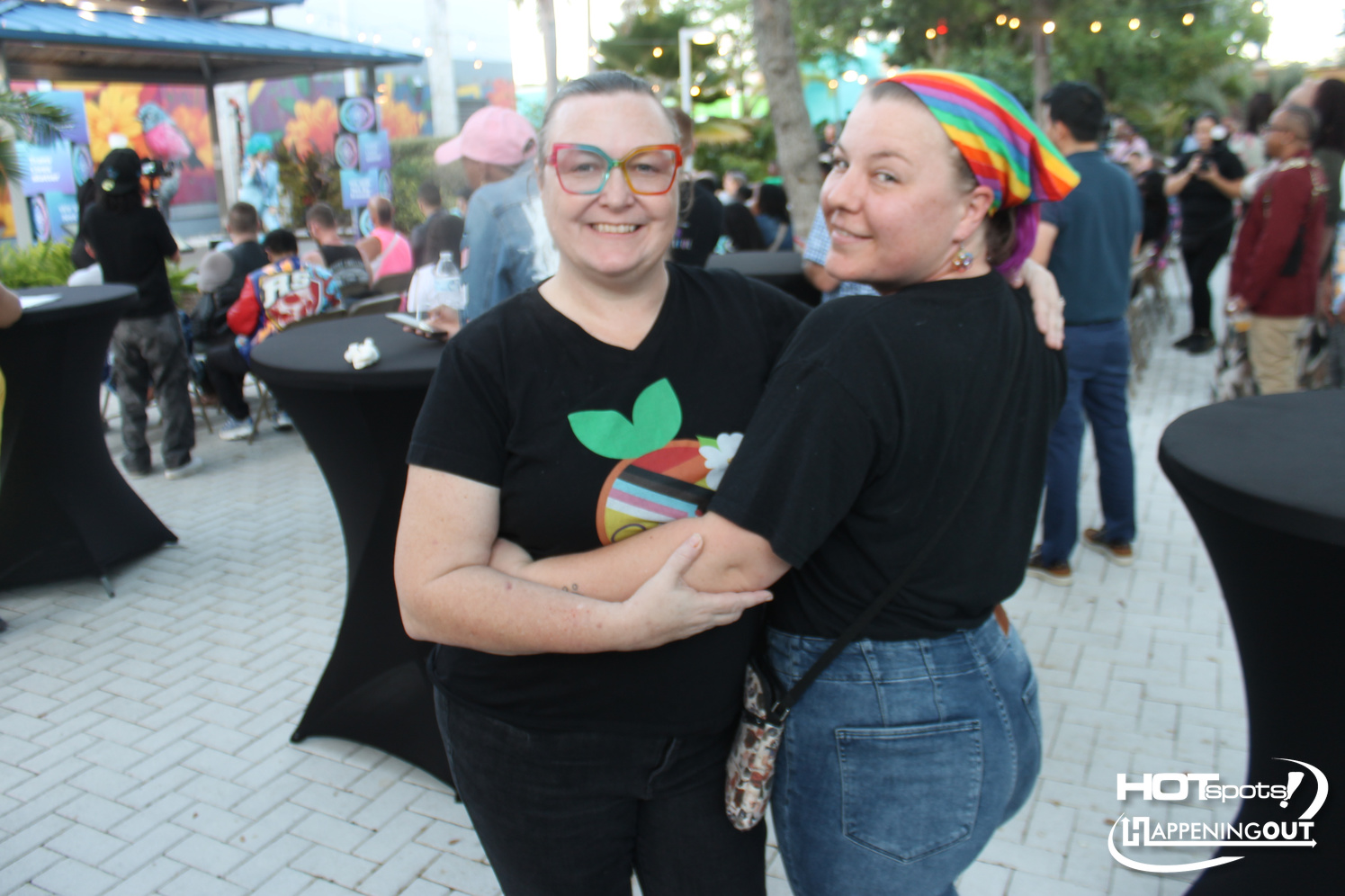 Two women at an outdoor festival hug and smile, one with colorful glasses and a rainbow headscarf two friends posing together at a sunny party with string lights in the background