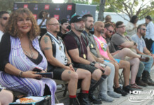 Group of people seated in a row at an outdoor event, watching a stage; includes a woman in a purple striped dress and several men with medals and tattoos.