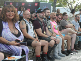 Group of people seated in a row at an outdoor event, watching a stage; includes a woman in a purple striped dress and several men with medals and tattoos.