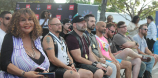 Group of people seated in a row at an outdoor event, watching a stage; includes a woman in a purple striped dress and several men with medals and tattoos.