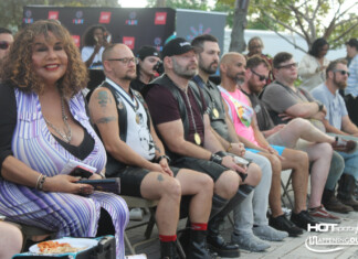 Group of people seated in a row at an outdoor event, watching a stage; includes a woman in a purple striped dress and several men with medals and tattoos.