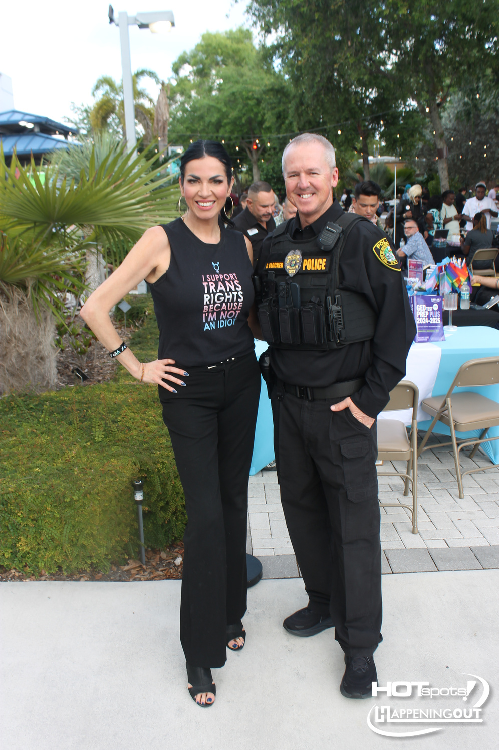 Smiling woman in a black sleeveless top with a transgender rights slogan stands next to a smiling police officer in tactical gear at an outdoor community event with tables and attendees in the background.