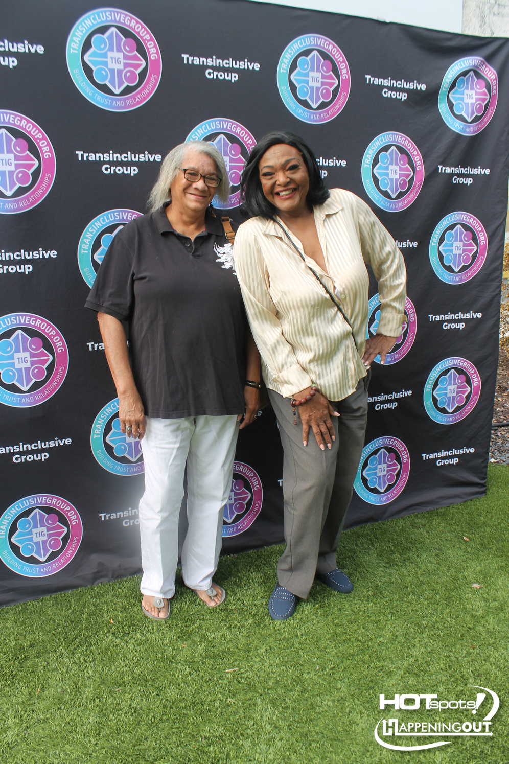 Two women standing on grass, smiling for the camera in front of a backdrop covered with circular Transinclusive Group logos; one wears a black polo and white pants, the other a beige striped blouse and gray pants.