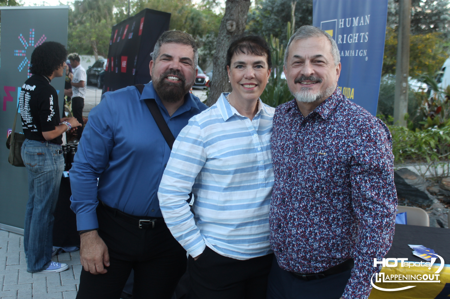 Three adults smiling at the camera at an outdoor event, with a blue Human Rights Campaign banner in the background.