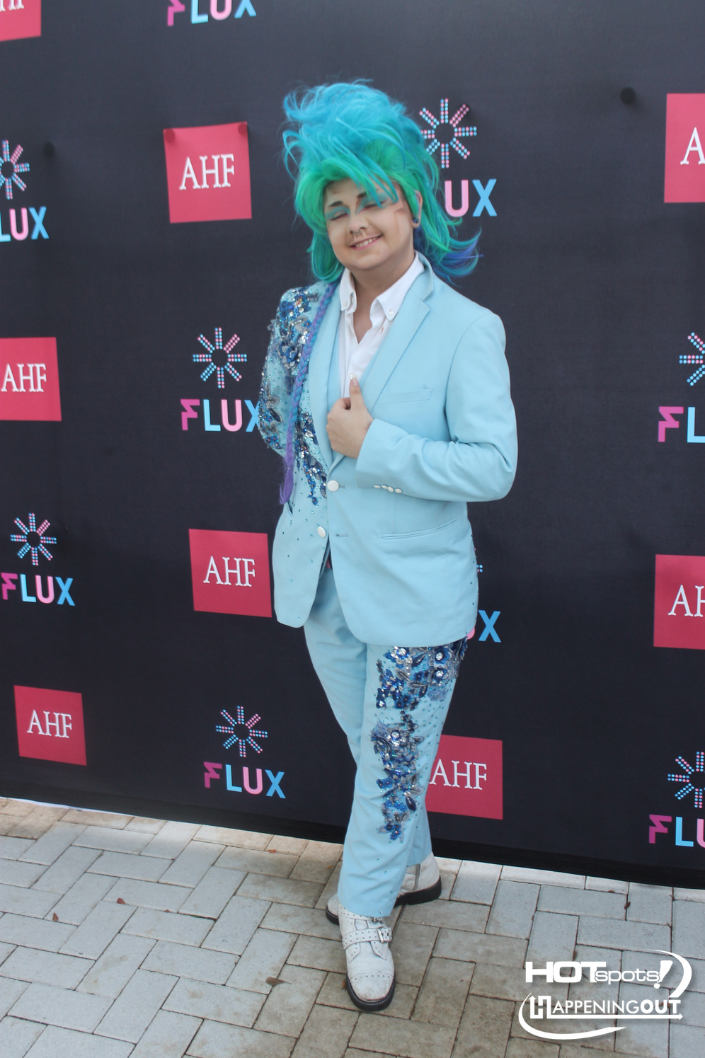 Person in a light blue suit with sequined embroidery, bright turquoise hair, posing at an event against a branded backdrop.