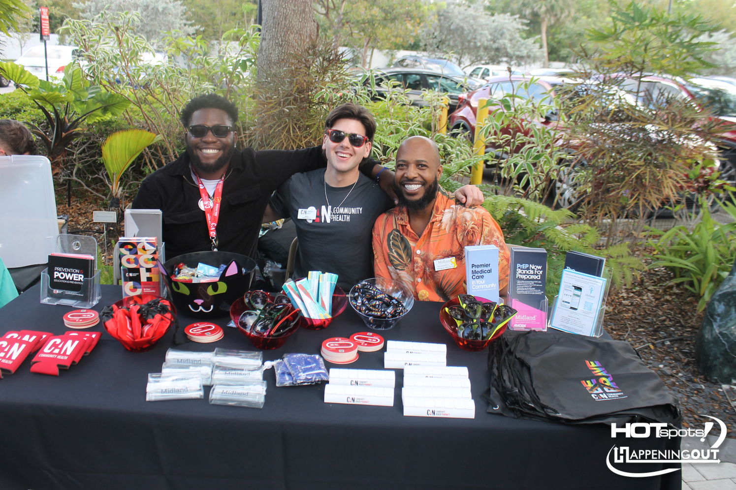 Three men at an outdoor health fair table with CN Community Health branding and various promotional items.