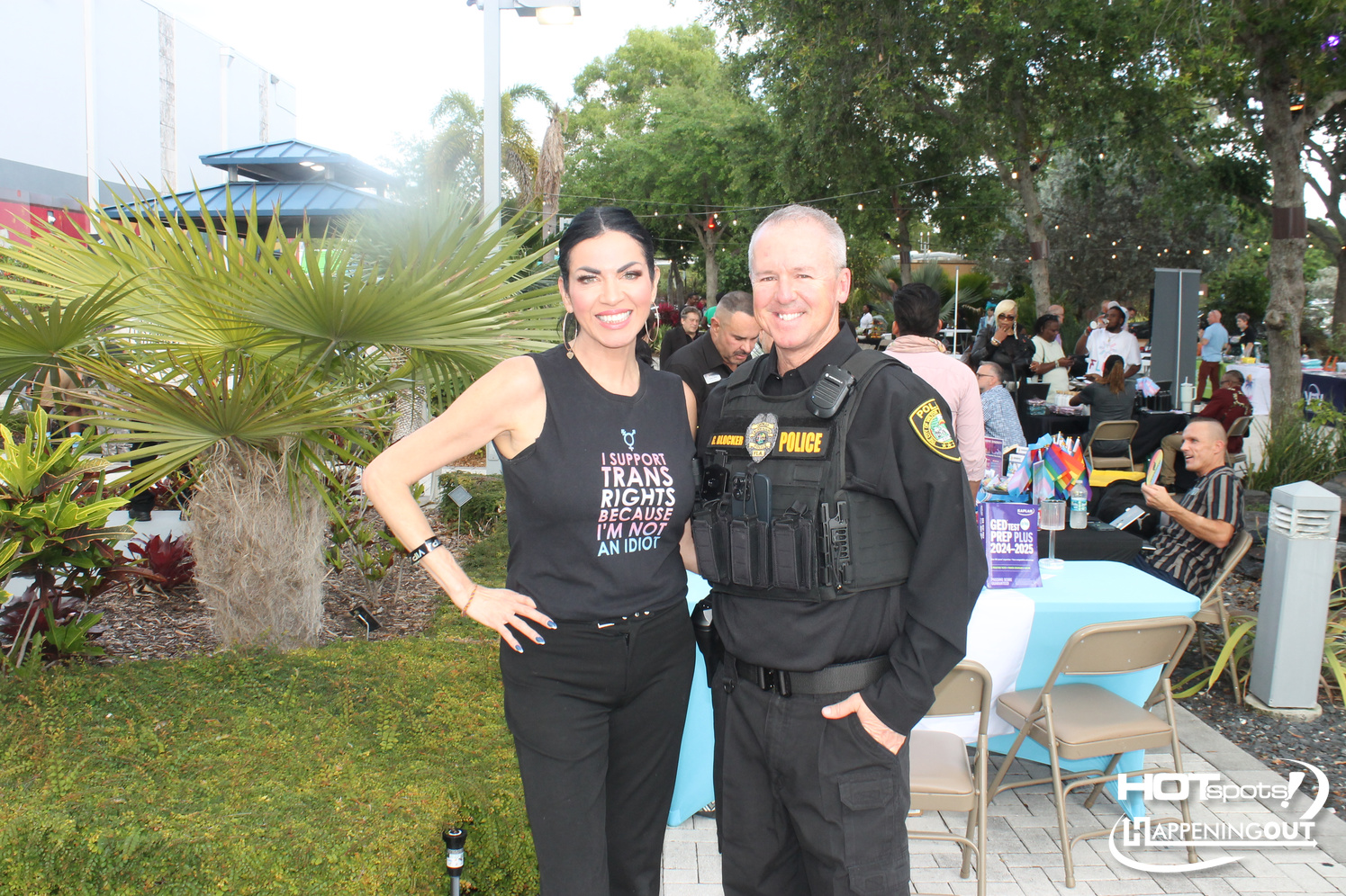 A smiling woman in a sleeveless black shirt standing next to a uniformed police officer at an outdoor community event.