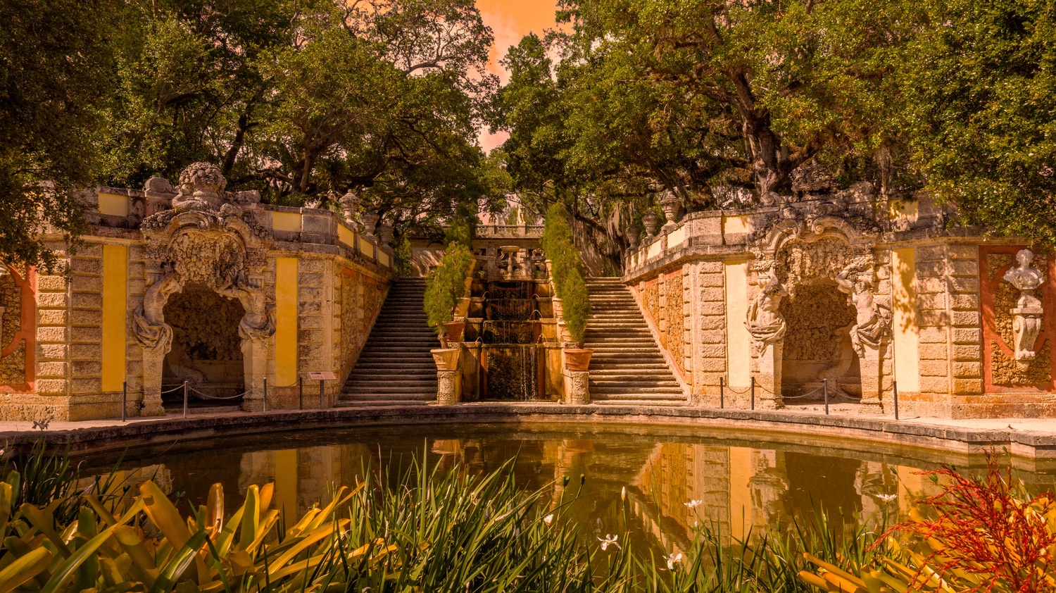 Symmetrical stone staircases flank a central fountain in a formal garden, with a reflective pond and lush trees overhead.