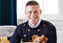 Smiling man in a navy sweater with small gold bee motifs, holding a small tan and white dog indoors.