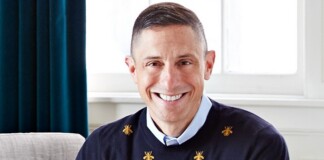 Smiling man in a navy sweater with small gold bee motifs, holding a small tan and white dog indoors.