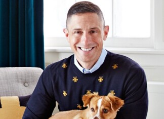 Joyful Output and a Joyless Outlook – Jonathan Adler on AIDS, survival, social media and why exuberance never seems as simple as it looks Smiling man in a navy sweater with small gold bee motifs, holding a small tan and white dog indoors.