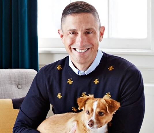 Smiling man in a navy sweater with small gold bee motifs, holding a small tan and white dog indoors.