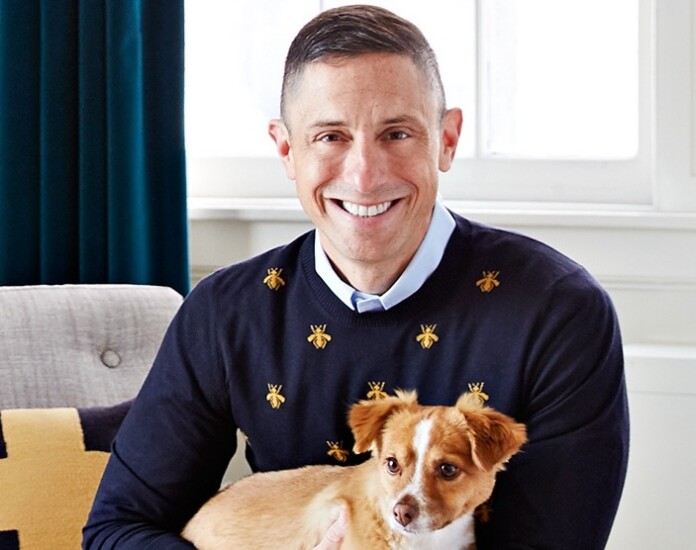 Smiling man in a navy sweater with small gold bee motifs, holding a small tan and white dog indoors.
