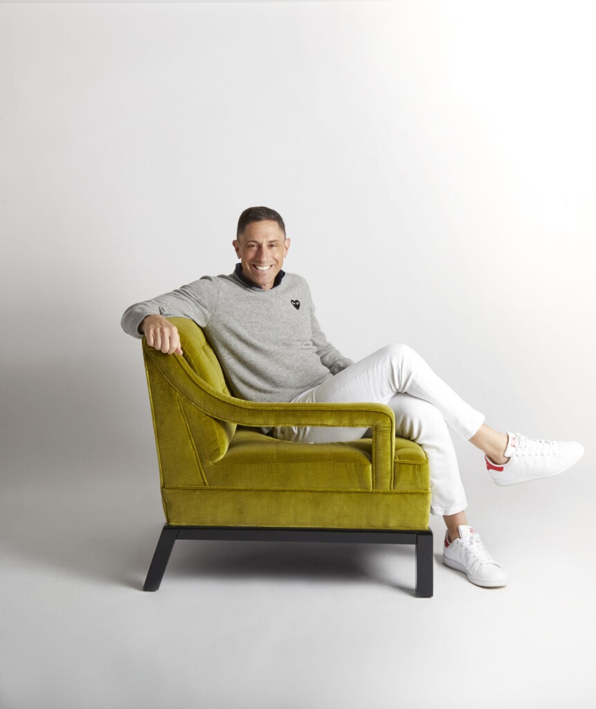 Smiling man in a gray sweater seated sideways in a mustard-yellow chair against a plain white background.