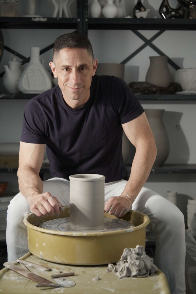 Man shaping clay on a pottery wheel in a studio, concentrating on his ceramic piece with tools nearby.
