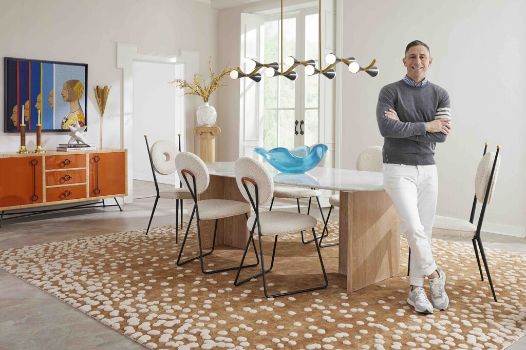 Man in a gray sweater and white pants stands beside a wooden dining island in a bright, stylish dining room with a dotted rug and modern chandelier.
