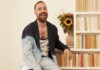 Man with a beard laughs while sitting beside a bookshelf with a bouquet of sunflowers in a vase behind him.