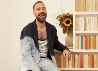 Man with a beard laughs while sitting beside a bookshelf with a bouquet of sunflowers in a vase behind him.